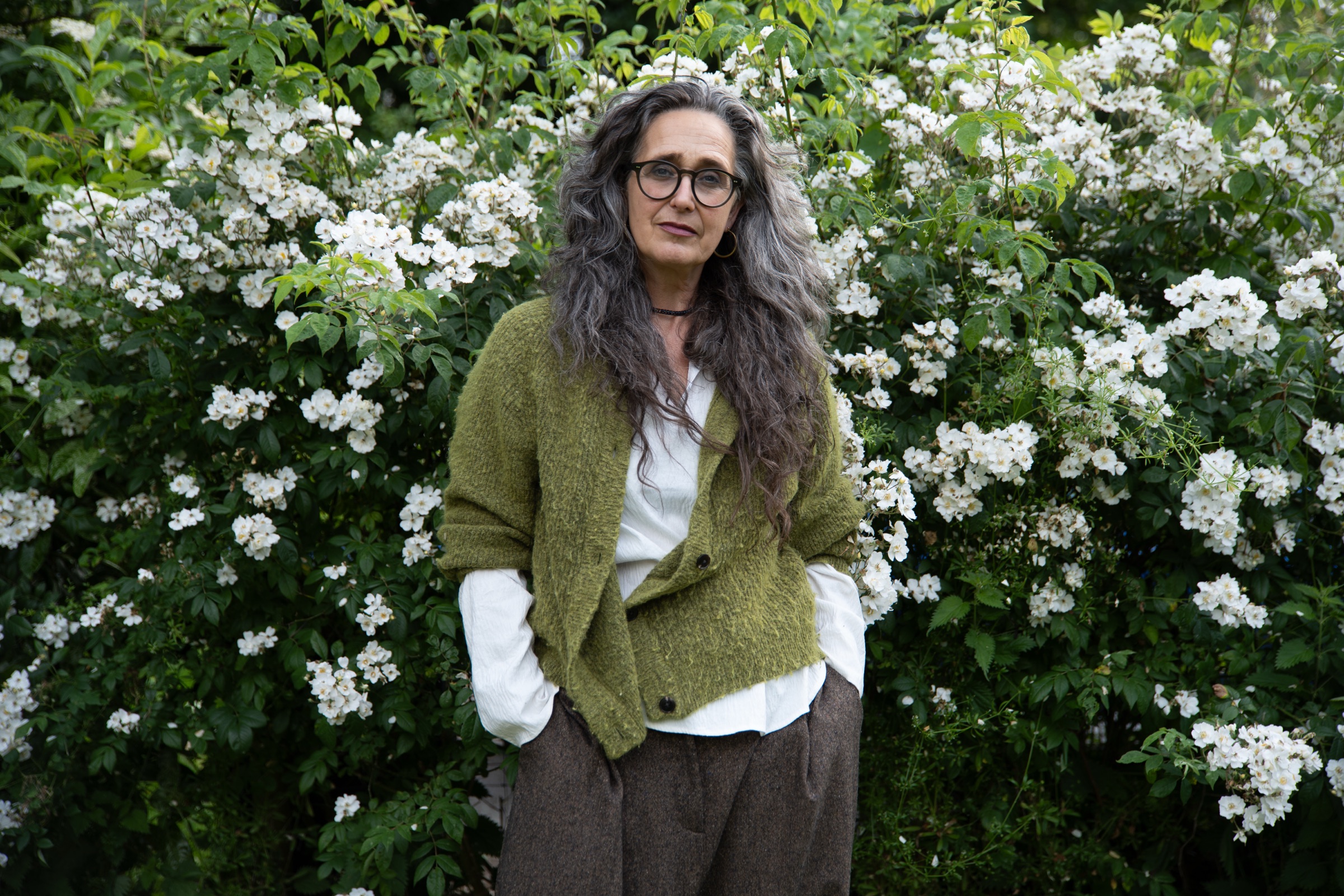 Christiane Churchill-Ley in a green cardigan standing before a bank of white rambling roses.